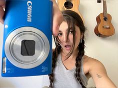 a woman holding up a camera in front of her face with guitars on the wall behind her