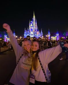 two women taking a selfie in front of the disney castle at night with their arms up