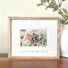 a white vase with a green plant in it next to a photo frame on a wooden table