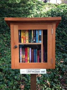 a wooden bookcase with many books on it in front of some bushes and trees