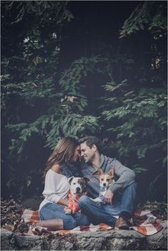 a man and woman sitting on a blanket with two dogs in front of some trees