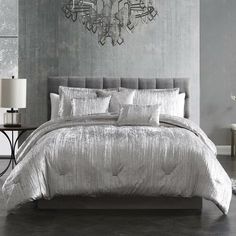 a bed with silver comforter and pillows in a gray room next to a lamp