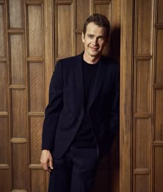 a man in a black suit leaning against a wooden wall with his hands on his hips