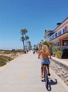 a woman riding a bike down a sidewalk next to palm trees and beachfront buildings