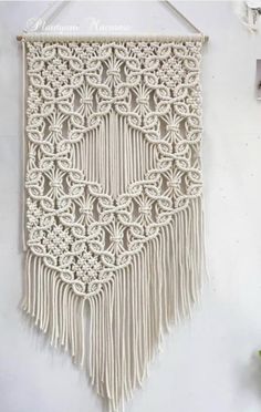 a white wall hanging with crochet and tassels on the bottom, next to a potted plant