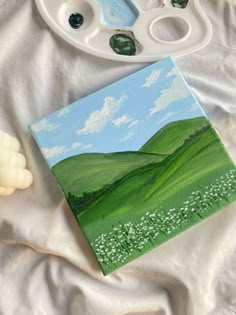 a painting is shown next to some paintbrushes on a white sheet with a green field and blue sky in the background