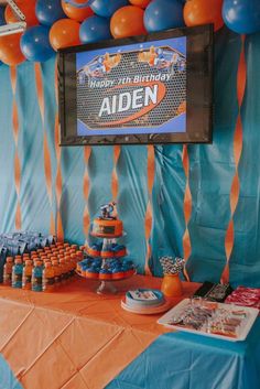 an orange and blue birthday party with balloons, streamers and a large screen on the wall