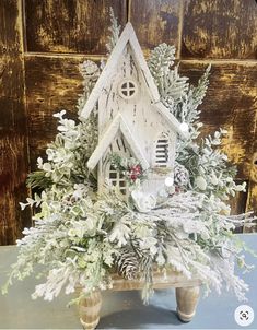 a white birdhouse surrounded by greenery and pine cones on a wooden table in front of a door