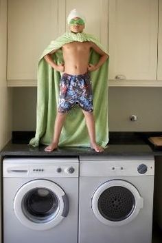 a kid standing on top of a dryer in front of a washing machine with a towel over his head