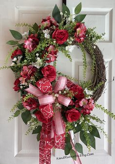 a wreath with red flowers and greenery hanging on a white front door decorated with pink ribbon