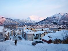 a person walking down a snow covered street next to houses in the distance with mountains in the background