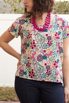 a woman wearing a multicolored floral top with a pink beaded necklace standing in front of a white fence