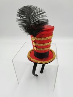 Nicky Bigs Novelties™ Deluxe Majorette Mini Top Hat Headband It's Circus time and you are the Ring Master. This satin Mini Ring Master hat is the perfect addition to your Circus themed party costume. Mini red top hat on a headband with black feather and gold braid embellishments. Top hat is approximately 5 inches tall, 5 inches in diameter, and attached to a plastic headband covered in black material. One size fits most. Handmade, assembled, and crafted by hand with assistance from hand driven fabric forming and hand driven sewing equipment. Themed Red Costume Hat, Adjustable Red Hat For Costume Party, Red Costume Hats And Headpieces For Carnival, Red Adjustable Hat For Costume Party, Novelty Mini Hats In Red For Gifts, Novelty Mini Red Hat For Gift, Themed Red Party Hat, Red Themed Costume Hats And Headpieces, Red Themed Hat For Costume Party