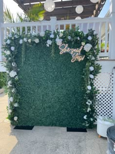 a green wall with white flowers and greenery on the top is surrounded by lights