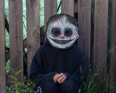 a person wearing a creepy mask sitting in front of a fence with grass and plants