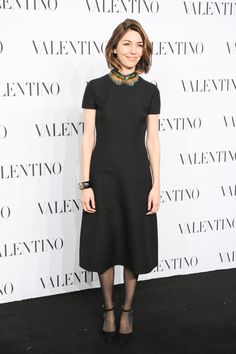 a woman standing in front of a wall wearing a black dress and high heeled shoes