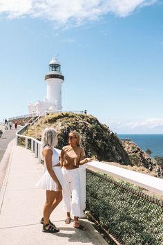 two women standing next to each other near the ocean with a lighthouse in the background