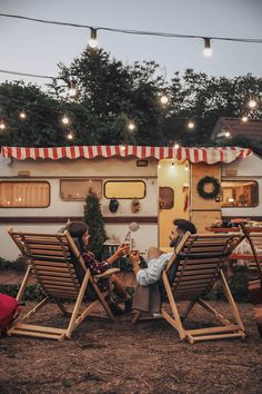 two people sitting on lawn chairs in front of a camper with lights strung over it