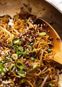 a wooden spoon is full of noodles and meat with sesame seeds on the side as it sits in a skillet