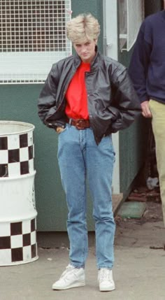 a woman standing in front of a building with her hands on her hips, wearing jeans and a leather jacket