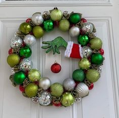 a christmas wreath on the front door decorated with green and red ornaments, including a handprinted frog ornament