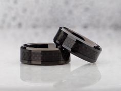 two wedding bands with black and gray carbon fiber inlays on each side, set against a white background