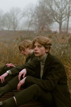 two young men sitting next to each other in front of tall grass and trees on a foggy day