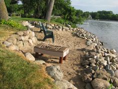 a bench sitting on the side of a river next to some rocks and grass with trees in the background