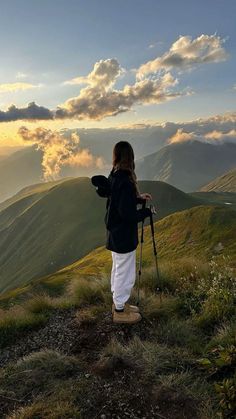 a woman standing on top of a lush green hillside under a cloudy sky with mountains in the background