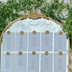 a wedding seating chart is displayed on a gold framed mirror with greenery around it