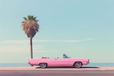 a pink convertible car parked on the side of the road next to a palm tree