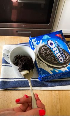 A spoon scooping chocolatey Oreo mug cake with a creamy center from a white mug, alongside a package of Oreo cookies on a striped kitchen towel.