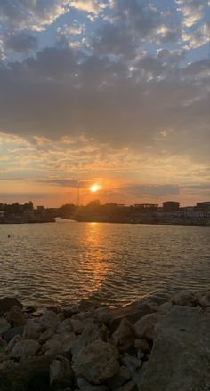 the sun is setting over water with rocks in front of it and buildings on the other side