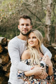 a man and woman hugging each other while standing in front of stacked logs with trees behind them