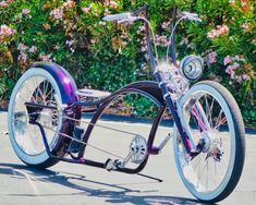 a purple and black bike parked on the side of a road next to flowers in front of a bush