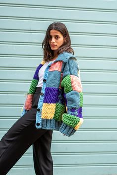 a woman wearing a multicolored knitted jacket and black pants standing in front of a blue garage door