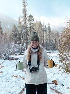 a woman standing in the snow with her camera and coffee mugs on her hands