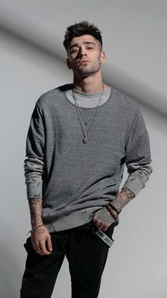 a young man with tattoos standing in front of a white wall wearing a grey sweater and black pants