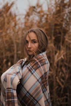 a woman wrapped up in a plaid blanket standing next to some tall grass and looking at the camera