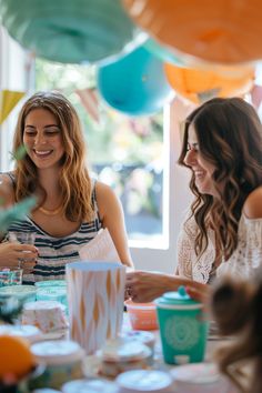 two women sitting at a table with cups and plates in front of them, laughing