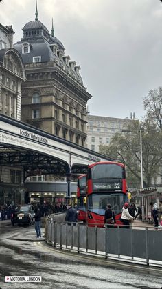 a red double decker bus parked in front of a train station with people standing on the sidewalk