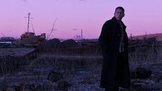 a man standing in the middle of a field at dusk with a ship in the background