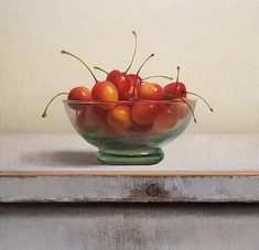 a green glass bowl filled with cherries on top of a wooden table next to a white wall