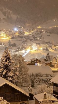 a snow covered village at night with lights on the buildings and trees in the foreground