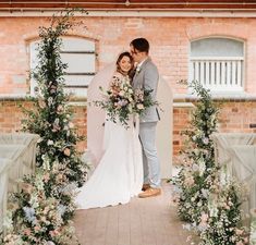 a bride and groom standing in front of an archway with flowers on the side, surrounded by greenery