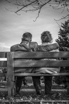 an elderly couple sitting on a bench looking at the trees and sky in black and white