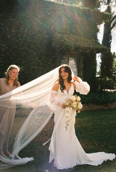 a woman in a wedding dress holding a veil over her head while another woman looks on