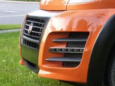 the front end of an orange car parked on grass next to a street and trees