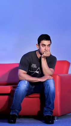 a man sitting on top of a red couch in front of a blue wall with his hand under his chin