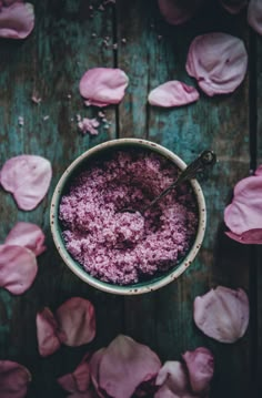 a bowl filled with pink sugar surrounded by petals on top of a wooden table next to a spoon